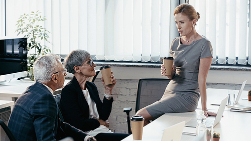 Manager holding coffee to go and talking to mature colleagues near gadgets in office
