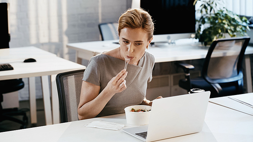 Businesswoman eating fresh salad near coffee to go and laptop in office