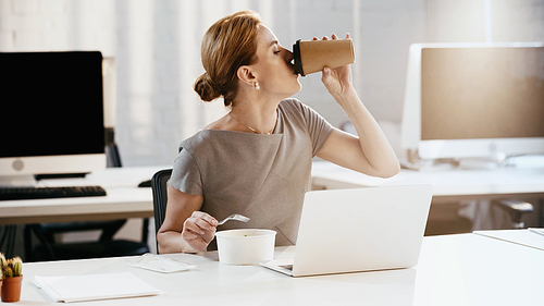 Side view of businesswoman drinking coffee to go near fresh salad and laptop in office