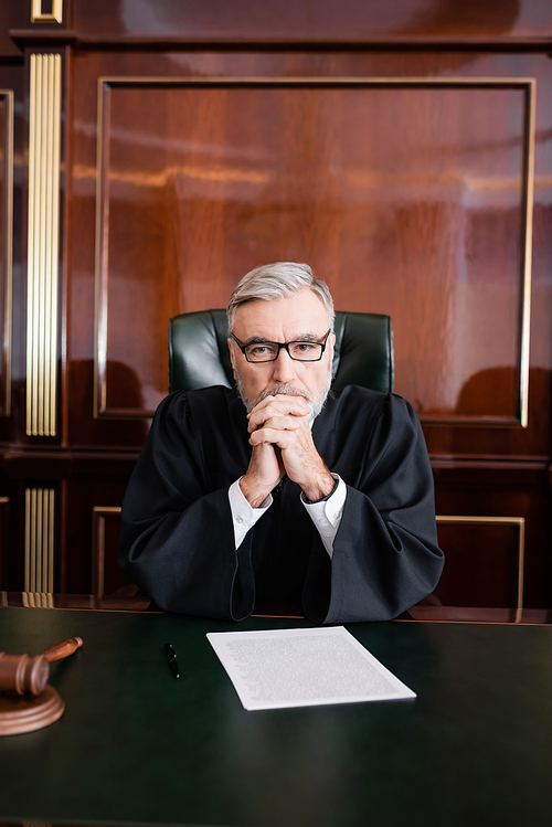 grey-haired judge in robe and eyeglasses sitting with clenched hands near lawsuit on desk