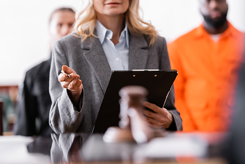 advocate with clipboard standing near accused african american man and bailiff on blurred background