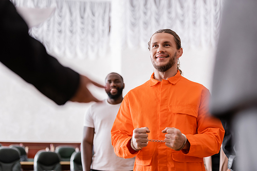 happy man in handcuffs and jail uniform near blurred jurors in courtroom