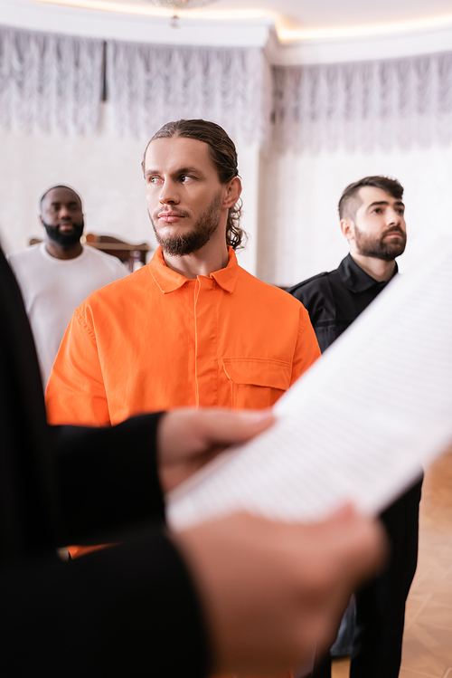 accused man in orange jail uniform standing near bailiff and prosecutor with paper on blurred foreground