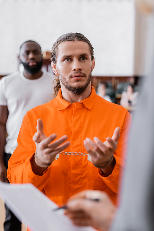 accused man in jail uniform and handcuffs gesturing near blurred prosecutor and african american juror in court