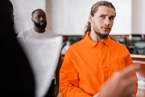 accused man in jail uniform looking at blurred prosecutor pointing with finger in courtroom