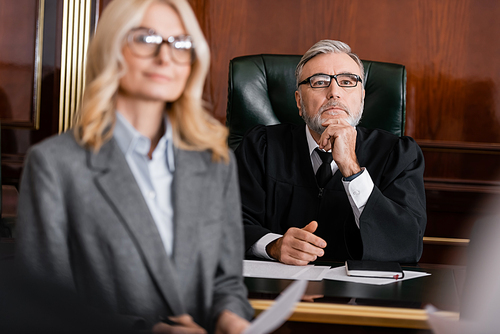 grey-haired judge in robe and eyeglasses near blurred advocate in courtroom