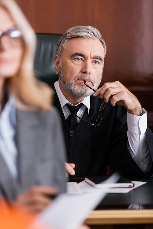 thoughtful judge holding eyeglasses near advocate on blurred foreground