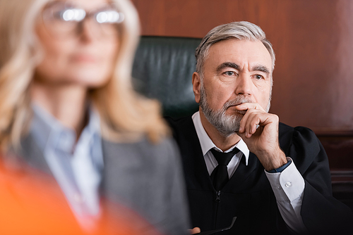 senior judge holding hand near face while thinking near advocate on blurred foreground