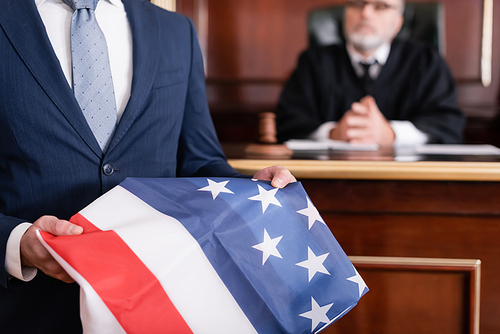 lawyer in suit holding usa flag in courtroom near senior judge on blurred background