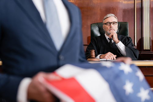 grey-haired judge  in court while attorney holding usa flag on blurred foreground