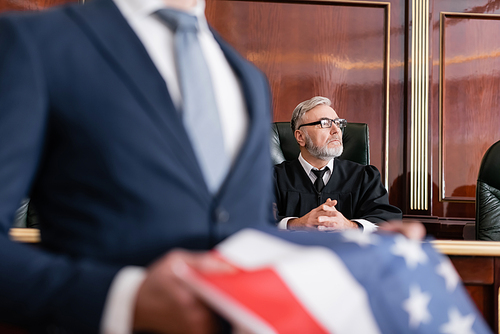 senior judge looking away in courtroom near attorney with usa flag on blurred foreground