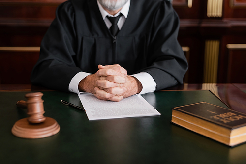 partial view of judge sitting with clenched hands near bible, gavel and lawsuit on desk