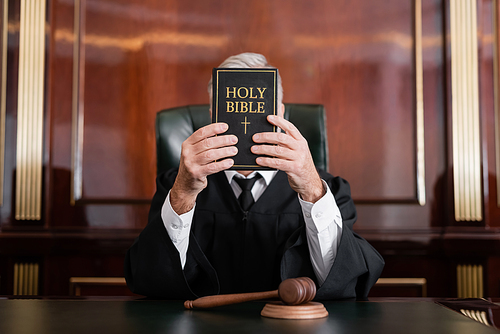 judge in mantle obscuring face with bible while sitting near gavel in courtroom