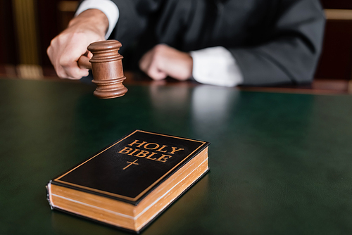 partial view of blurred judge holding gavel near holy bible on desk