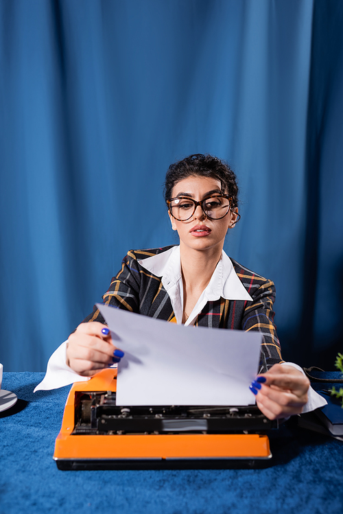 journalist in vintage style clothes holding empty paper near typewriter on blue background