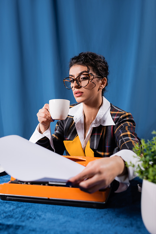 vintage style newswoman with coffee cup holding empty paper near typewriter on blue background
