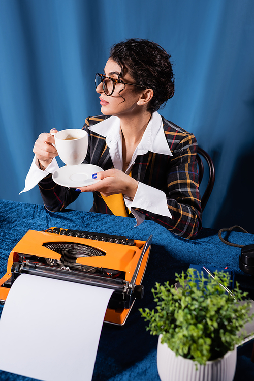 stylish journalist sitting with coffee cup near vintage typewriter on blue background