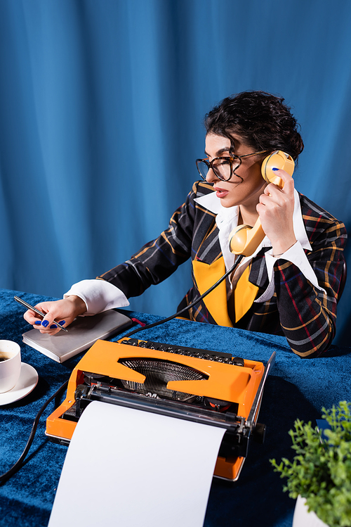 brunette newswoman in vintage blazer holding pen while talking on telephone on blue background