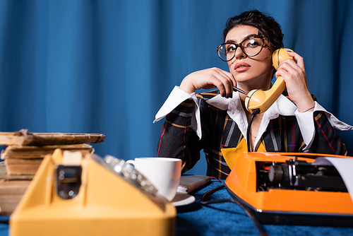 thoughtful newswoman talking on blurred telephone near typewriter on blue background