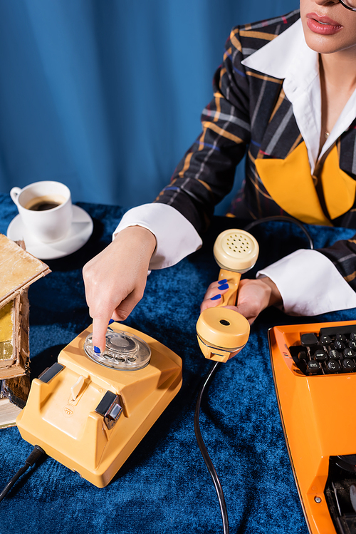 partial view of stylish woman dialing phone number near typewriter and coffee cup on blue background
