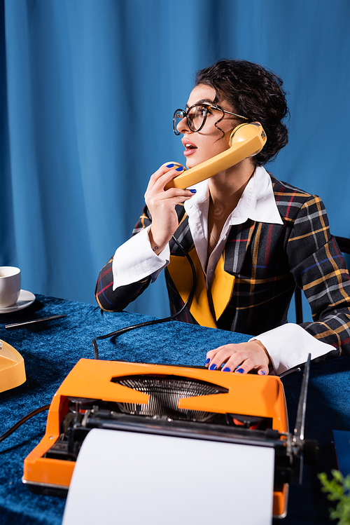 retro style journalist talking on telephone near typewriter on blue background with drapery