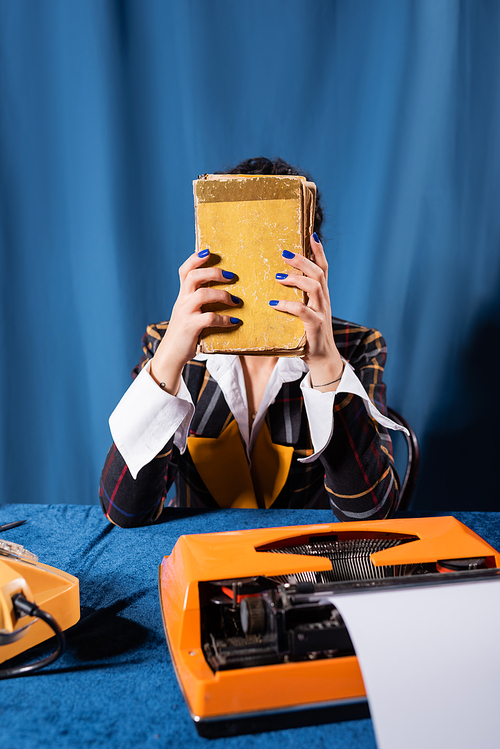 newswoman obscuring face with book near vintage typewriter on blue background