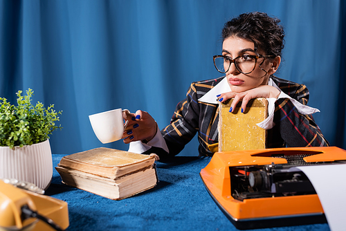 pensive woman sitting with coffee cup and book near vintage typewriter on blue background