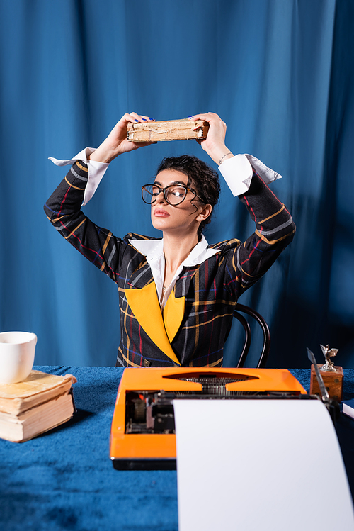 retro style newswoman meditating with book above head near typewriter on blue background