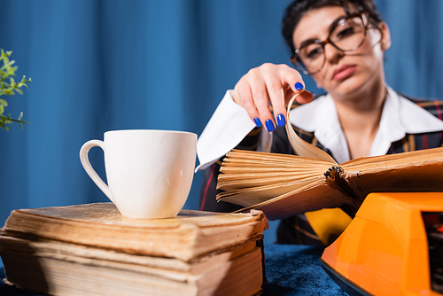 coffee cup near blurred newswoman in eyeglasses reading vintage book on blue background