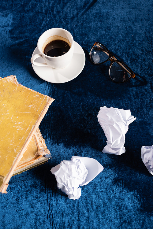top view of tattered books, crumpled paper, eyeglasses and coffee cup on blue velour tablecloth
