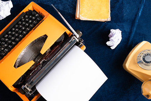 top view of vintage typewriter with blank paper, telephone and old book on blue tablecloth