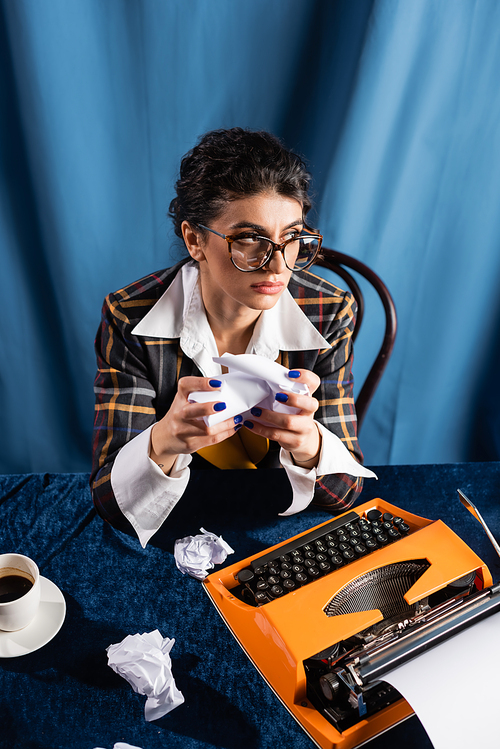 thoughtful newswoman with crupled paper looking away near vintage typewriter on blue background