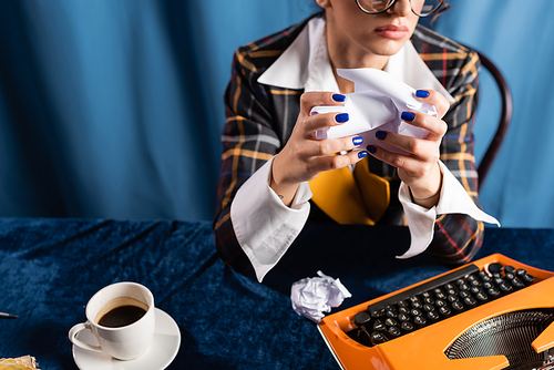 partial view of stylish woman with crumpled paper near vintage typewriter and and coffee cup on blue background