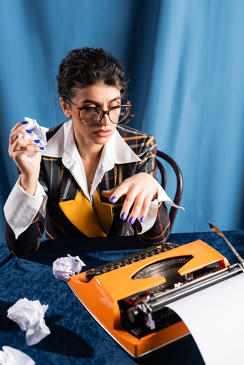 vintage style journalist holding crumpled paper near typewriter on blue background