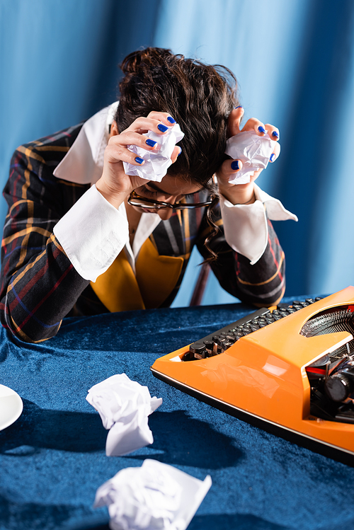 depressed journalist sitting with bowed head and crumpled paper near vintage typewriter on blue background