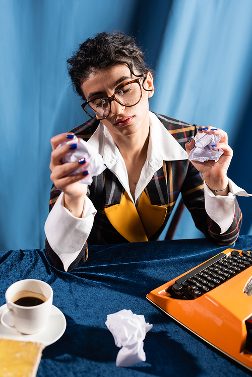 upset and exhausted journalist holding crumpled paper near typewriter on blue background