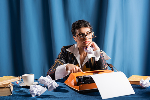 thoughtful newswoman looking away near crumpled paper and vintage typewriter on blue background