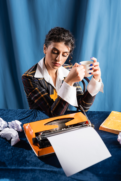 retro style journalist with coffee cup sitting near typewriter and crumpled paper on blue background