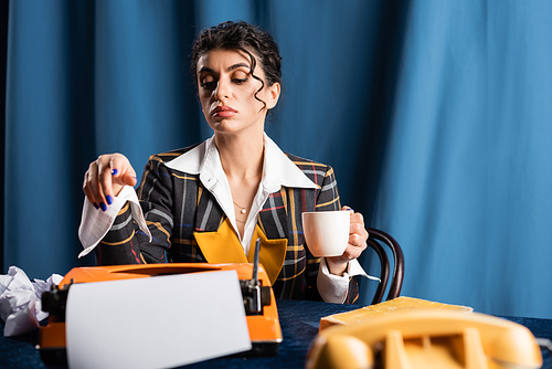 stylish journalist with coffee cup near vintage typewriter on blue background
