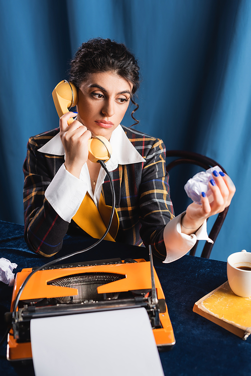 pensive newswoman with crumpled paper talking on telephone near typewriter on blue background