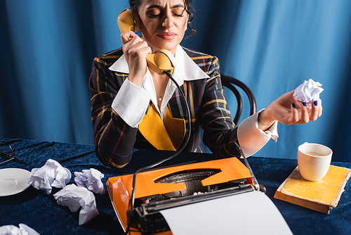 displeased woman with closed eyes holding crumpled paper and talking on telephone on blue background