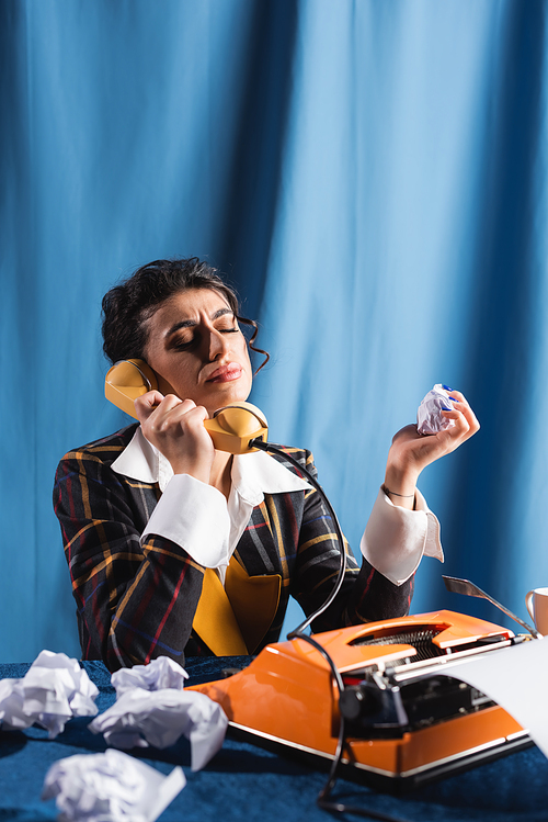 upset woman holding crumpled paper while talking on telephone near typewriter on blue background