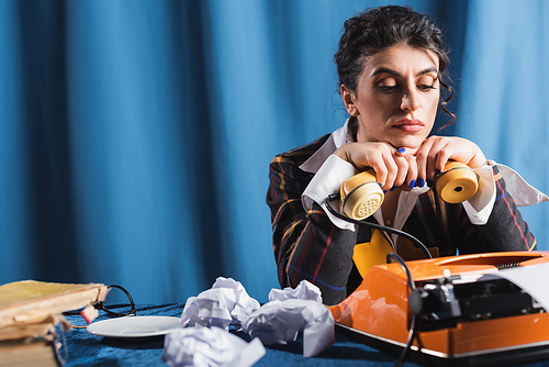 upset newswoman sitting with handset near vintage typewriter and crumpled paper on blue background