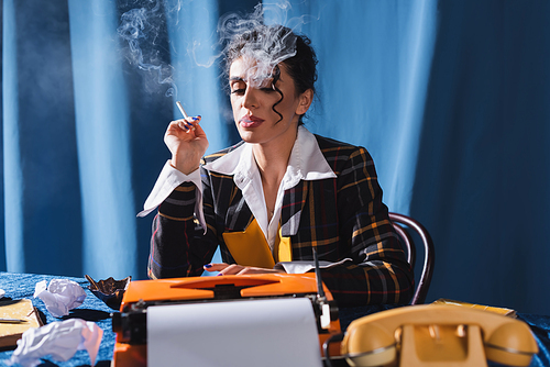 stylish woman in vintage style clothes smoking near typewriter and crumpled paper on blue background