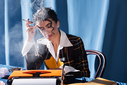 stylish woman typing on vintage typewriter and smoking on blue background