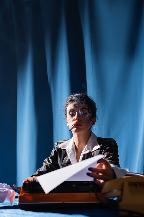 smoking vintage style woman holding paper near typewriter on blue background with drapery