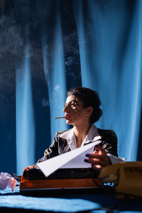 stylish woman holding paper near blurred typewriter while smoking on background with blue drapery