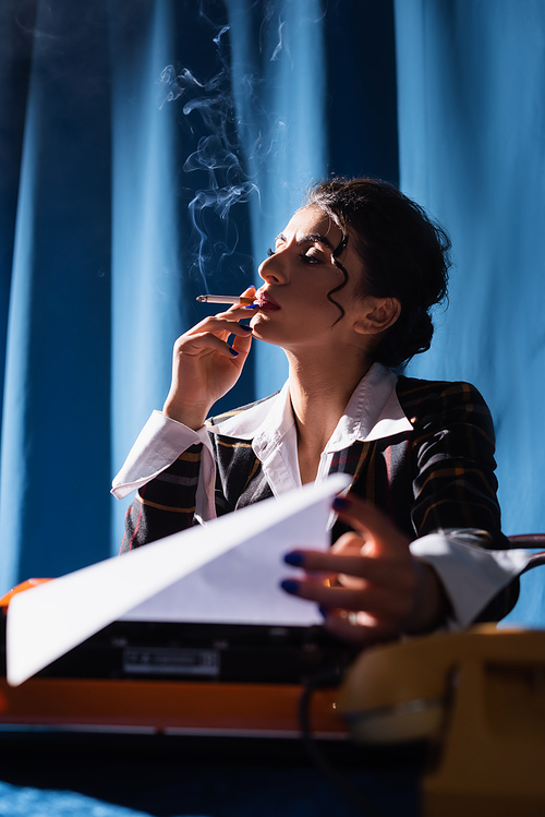 stylish woman in retro style clothes smoking near typewriter on blue background with drape