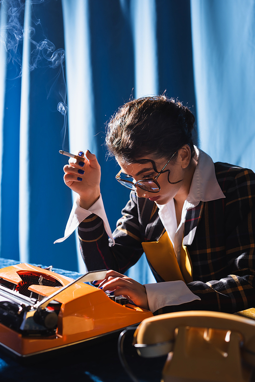 stylish newswoman in eyeglasses holding cigarette while working at typewriter on blue background