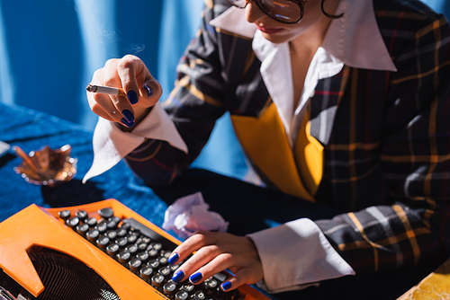 cropped view of blurred woman with cigarette typing on vintage typewriter on blue background